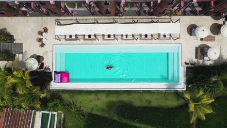 Female-swimming-looking-up-at-the-sky-at-Shore-Amora-Canggu-Hotel-pool-in-Bali,-Indonesia,-Aerial-rising-reveal-shot