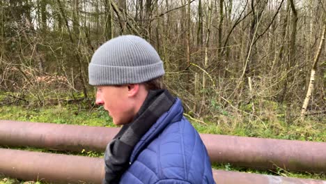 Slow-motion-shot-of-thoughtful-young-man-with-hat-walking-along-rural-forest-path-during-autumn-with-leafless-trees-in-background