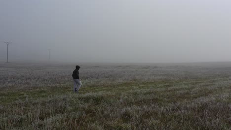 Lonely-Man-Walking-on-Meadow-on-Foggy-Morning,-Tracking-Drone-Aerial-View