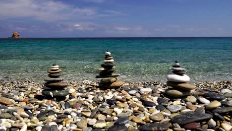 Three-Zen-stacked-rock-towers-on-a-beach-with-crystal-clear-water-in-the-background-in-Sardinia,-Italy