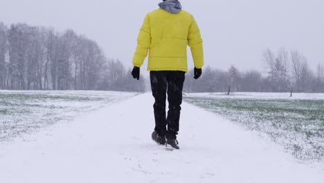 Rear-shot-of-man-in-yellow-winter-jacket-walking-on-snowy-path-during-a-cloudy-cold-winter-day