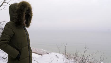 Young-woman-in-green-winter-clothes-standing-on-the-steep-dunes-along-the-sandy-shore-of-the-Baltic-sea-beach,-old-military-building-ruins,-medium-tracking-shot