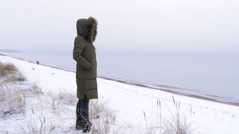 Young-woman-in-green-winter-clothes-stands-on-the-sandy-shore-of-the-Baltic-sea-beach,-snowy-grass,-medium-jib-shot-up