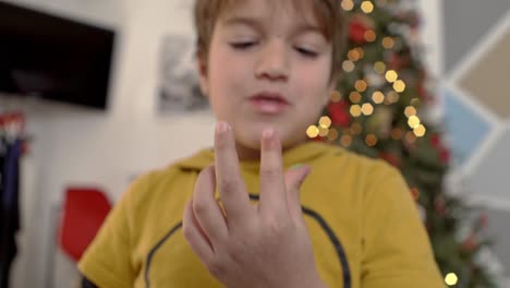 Caucasian-kid-licking-sugar-icing-from-his-fingers,-Christmas-tree-on-the-blurry-background-4k