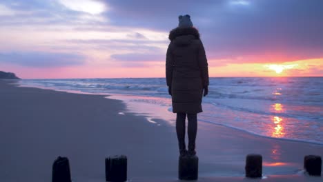 Young-woman-in-winter-clothes-standing-on-old-wooden-pier,-sandy-shore-of-the-Baltic-sea-beach,-watching-romantic-sunset,-medium-tracking-shot-right