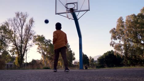 Ground-level,-back-view-of-caucasian-boy-taking-a-basketball-shot,-at-basket-hoop,-located-in-an-alley-amongst-trees