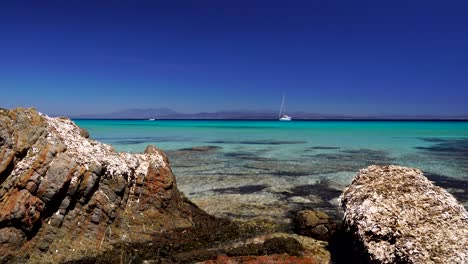 Die-Kamera-Zoomt-Zwischen-Zwei-Felsen-An-Einem-Wunderschönen-Strand-Mit-Einem-Segelboot-Im-Hintergrund