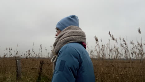 Tracking-shot-of-beautiful-young-woman-with-scarf-and-beanie-walking-between-agricultural-fields-outdoors-during-cloudy-day