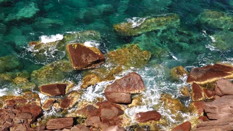 Die-Kamera-Blickt-Auf-Eine-Felsige-Küste-Mit-Roten-Felsen,-Kristallklarem-Wasser-Und-Sanften-Wellen