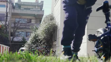 Ground-view-of-caucasian-kid-pushing-his-bicycle,-grass-in-the-blurry-foreground-120fps