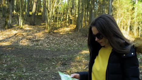 Young-caucasian-woman-with-brown-hair-and-dark-sunglasses-walking-in-the-wood,-map-in-hand,-beautiful-sunny-spring-day,-tracking-jib-shot-from-ground-up