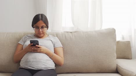 Eine-Junge-Schwangere-Latinx,-Die-Sich-Mit-Einem-Smartphone-Auf-Dem-Sofa-Entspannt