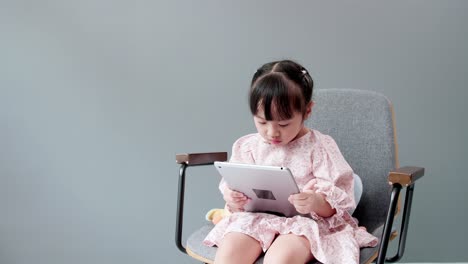 Little-asian-girl-sitting-on-chair,-swing-legs,-studying-online-e-learning-system-on-tablet,-paying-attention-to-online-video