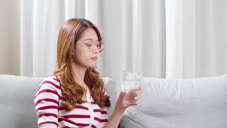 Mujer-Joven-Asiática-Sentada-En-El-Sofá-Bebiendo-Agua-Para-Relajarse-Durante-Un-Fuerte-Dolor-De-Cabeza