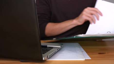 Male-Office-Worker-Looking-Through-Files-In-Green-Folder-With-Laptop-And-Papers-On-Wooden-Office-Table