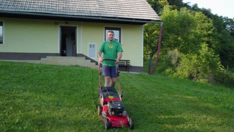 Happy-man-in-blue-shorts-and-green-shirt-mowing-green-lawn-in-front-of-his-house