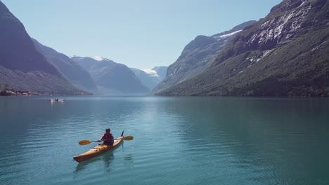 Woman-kayaking-turquoise-Lovatnet-lake-during-sunny-summer-day---Backward-moving-aerial-close-to-water-with-kayak-moving-towards-camera---Beautiful-Norway-mountain-landscape-with-glacier-in-background