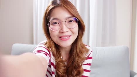 The-portrait-of-a-selfie-Asian-woman-with-eyeglasses-is-smiling-with-laughter-being-positive-emotions-on-video-call-online-sitting-on-the-sofa-in-the-living-room