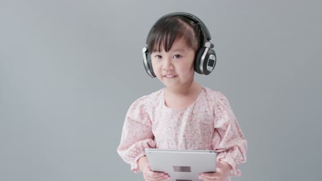 Little-Asian-girl-wearing-headphones-holding-tablet,-listening-to-music-and-dancing-happily