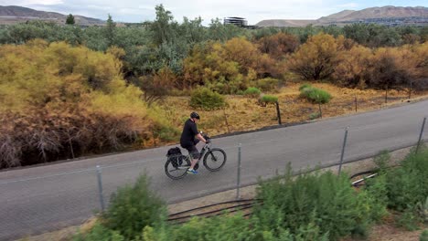 Mature-man-cycling-along-a-paved-nature-trail-with-sky-and-mountains-in-the-background---aerial-tracking-view