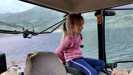 Farmers-cute-young-daughter-sitting-inside-tractor-close-to-Hardangerfjord-sea---Eager-to-learn-and-start-driving-tractor---Handheld-inside-tractor-cabin---Norway