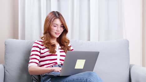 Young-Asian-woman-sitting-on-sofa-using-notebook-to-shop-online