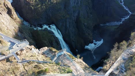 Spectacular-viewpoint-with-person-standing-on-the-edge-of-Vøringsfossen-waterfall-cliff---Waterfall-faalling-down-in-deep-canyon-below---Aerial-showing-walkway-and-viewpoints-over-waterfall---Norway