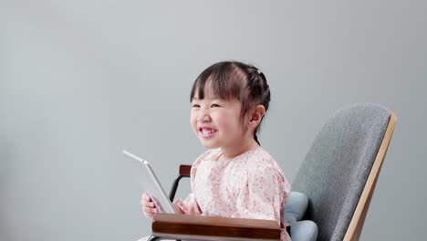 Asian-child-with-fringe-in-hair-sitting-on-chair-laughing-while-using-digital-tablet