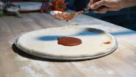 Masa-De-Pizza-En-Una-Bandeja-De-Acero-Sobre-Un-Mostrador-De-Madera-Mientras-Un-Chef-Experimentado-Le-Pone-Salsa-Roja-Con-Un-ácido
