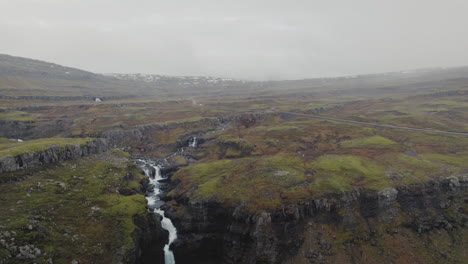 Aerial-dolly-in-over-river,-waterfall-towards-road-through-mountain-steppes-of-Iceland