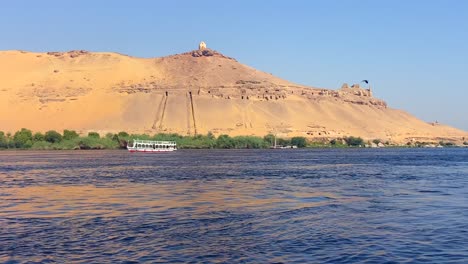4K-Aufnahmen-Von-Motorbooten,-Die-Eine-Wüstenlandschaft-Mit-Alten-ägyptischen-Gräbern-Am-Ufer-Des-Nils-überqueren