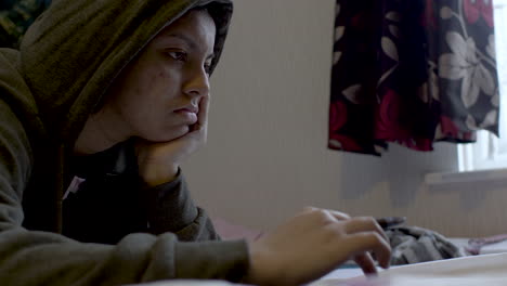 A-close-up-shot-of-a-young-female-high-School-pupil-dressed-in-a-hoodie-sitting-on-her-bed,-completing-her-online-homework-assignment-but-extremely-bored-as-the-current-subject-does-not-interest-her