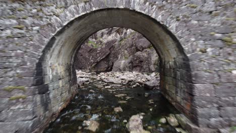 Flying-through-arch-of-old-traditional-stone-bridge-at-road-fv569-besides-Hesjedalsfossen-waterfall---Woman-swimming-in-pond-on-other-side---western-Norway