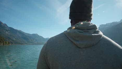 Strong-African-American-man-standing-quietly-in-deep-thought-as-ducks-pass-by-across-beautiful-lake-in-Interlaken-Switzerland,-Europe