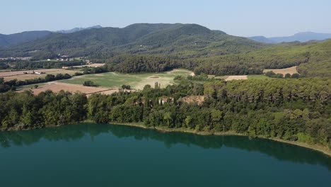 Aerial-view-over-a-beautiful-landscape-with-a-natural-lake-surrounded-by-woods,-and-farmlands-in-the-Spanish-mountains