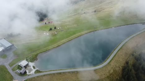 Drone-looks-down-on-clouds-floating-over-a-beautiful-small-reservoir-on-the-mountain-Asitz-Kogel-in-Austria,-Leogang-Saalbach