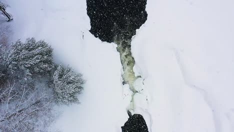 Fliegen-über-Einen-Kleinen-Wasserfall-Auf-Einem-Zugefrorenen-Fluss-Während-Eines-Schneesturms-In-Zeitlupe-Von-Oben-Nach-Unten