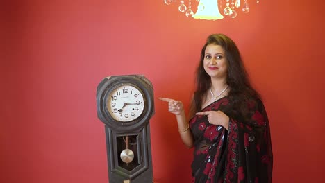 Eine-Indische-Frau-In-Schwarzem-Saree,-Die-Neben-Einer-Alten-Vintage-Uhr-In-Einem-Isolierten-Roten-Hintergrund-Mit-Goldenem-Licht-An-Der-Spitze-Steht