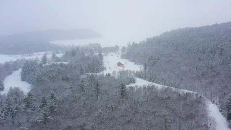 Flying-high-above-a-snow-covered-forest-during-a-blizzard-with-a-barn-in-the-distance-AERIAL