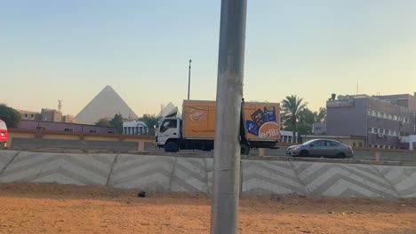 Side-view,-vehicles-drive-on-road-in-Egypt,-Great-Pyramids-of-Giza-in-background
