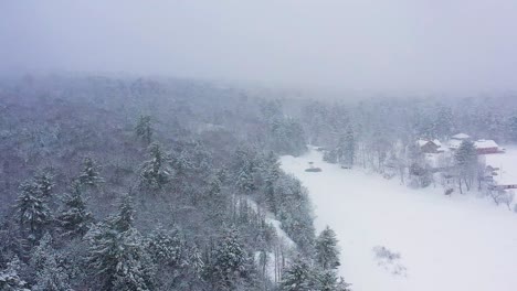 Aerial-orbit-above-a-forest-and-frozen-river-with-camp-along-its-shore-during-a-snow-storm-SLOW-MOTION