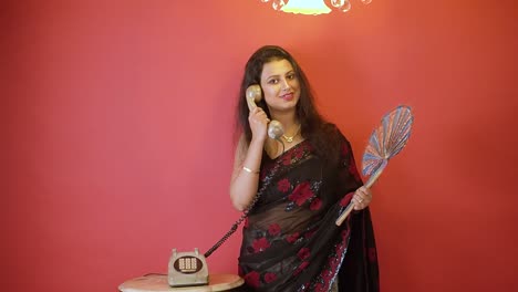 Eine-Indische-Frau-In-Schwarzem-Sari,-Die-Steht-Und-Im-Festnetztelefon-Mit-Einem-Handfächer-In-Einem-Isolierten-Roten-Hintergrund-Mit-Goldenem-Licht-Oben-Spricht