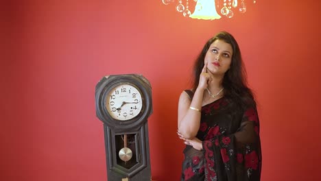 An-Indian-woman-in-black-saree-thinking-and-standing-next-to-a-old-vintage-clock-in-an-isolated-red-background-with-golden-light-on-top