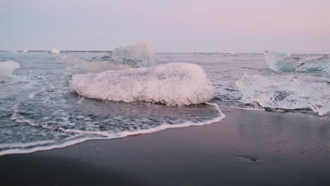 Handheld-pan-of-waves-washing-around-chunks-of-ice-on-Diamond-Beach,-Iceland