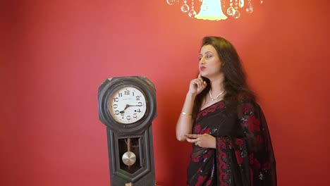 An-Indian-woman-in-black-saree-giving-thinking-expression-and-standing-next-to-a-old-vintage-clock-in-an-isolated-red-background-with-golden-light-on-top