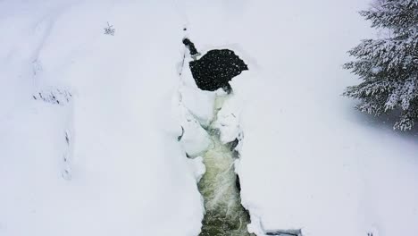 Während-Einer-Schneesturm-Zeitlupenantenne-Von-Einem-Kleinen-Wasserfall-Auf-Einem-Zugefrorenen-Fluss-Wegfliegen