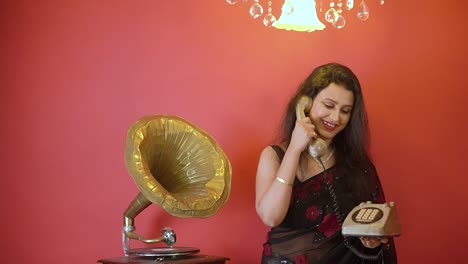 An-Indian-woman-in-black-saree-talking-in-telephone-catching-in-hand,-standing-next-to-gramophone-in-an-isolated-red-background-with-golden-light-on-top