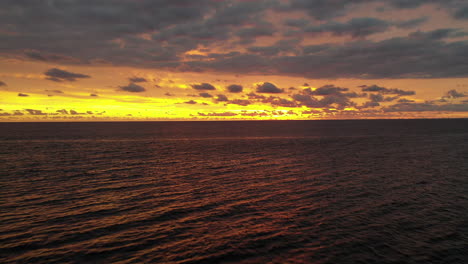 Luftflug-Rückwärts-über-Ruhiges-Meer-Mit-Einem-Dramatischen,-Hellgelben,-Orangefarbenen-Und-Violetten-Sonnenuntergang
