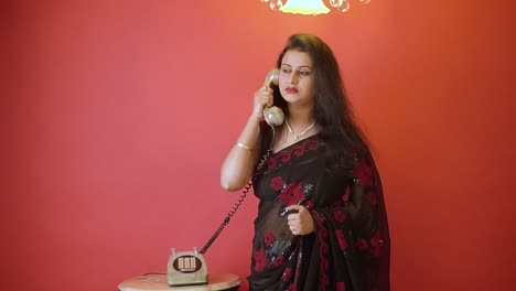 Eine-Indische-Frau-In-Schwarzem-Sari,-Die-Steht-Und-Im-Festnetztelefon-Mit-Einem-Lächeln-Auf-Einem-Isolierten-Roten-Hintergrund-Mit-Goldenem-Licht-Oben-Spricht