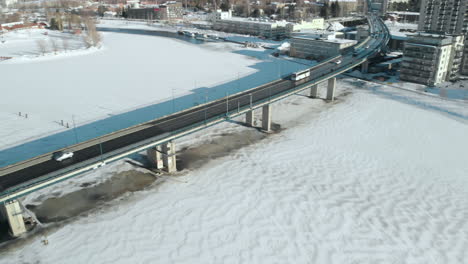 Aerial,-pan,-drone-shot,-above-Pielisjoki,-of-cars,-a-bus-and-traffic-on-Suvantosilta-bridge,-on-a-sunny,-winter-day,-in-Joensuu,-North-Karelia,-Finland